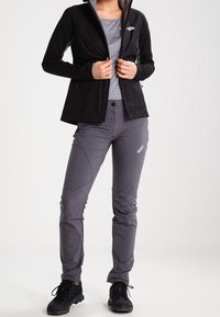 Černá zipová bunda, šedé tričko s dlouhým rukávem, šedé kalhoty se švy a logem, černé boty. Tkanina vypadá hladce a lehce.