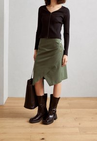 Haut à manches longues noir à côtes, jupe enveloppante vert olive avec des accents de boutons, bottes noires jusqu'aux genoux et un sac fourre-tout texturé noir.