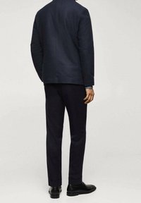 Man die zich omgekeerd bevindt, draagt een donkerblauw pak met bijpassende broek en zwarte dresschoenen, tegen een effen lichte achtergrond.