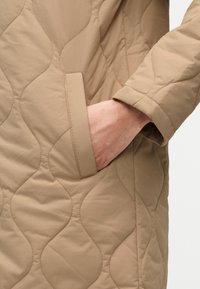 Μπεζ καπιτονέ παλτό με διακριτικό κυματιστό μοτίβο, με πλαϊνή τσέπη και απαλό ύφασμα. Κοντινή λήψη του χεριού που ξεκουράζεται στην τσέπη.