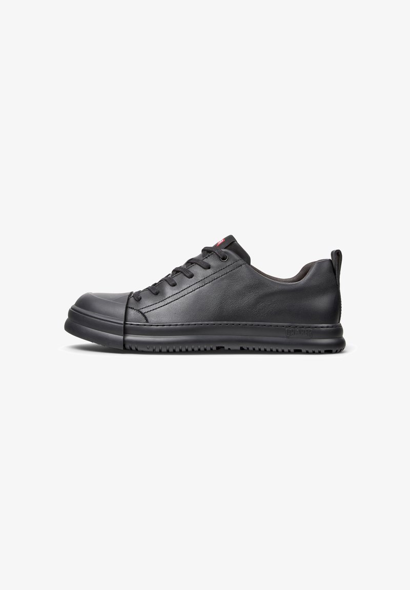 Zapatilla negra de cuero con textura suave, punta redondeada, cinco ojales y suela de goma gruesa. Diseño minimalista con un logotipo sutil en el lado.