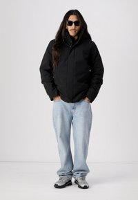 Svart isolert jakke med høy krage, blå dongeribukser og grå sneakers. Langt hår og solbriller fullfører looken.