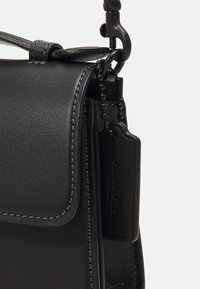 Bolso de hombro de cuero negro con cierre de solapa, que presenta costuras en contraste y una etiqueta con marca en relieve. Correa de eslabones de cadena.