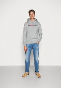 Šedá mikina s kapucňou, šnúrkami a predným vreckom, s čiernym nápisom loga. Svetlomodré džínsy a hnedé tenisky s bielymi detailmi.