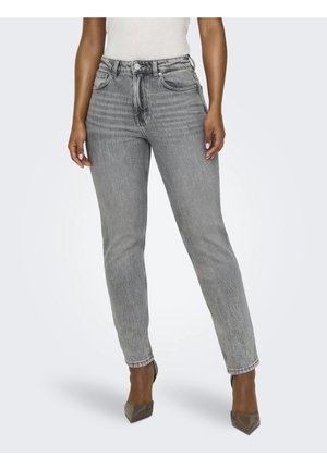 Person, die graue Jeans mit hohem Bund und taupefarbene spitze Absatzschuhe trägt, steht mit entspannten Händen an den Seiten vor einem einfarbigen Hintergrund.