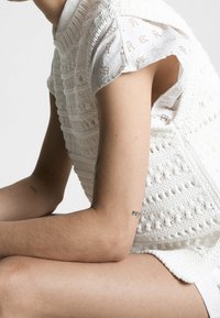 Top senza maniche bianco lavorato a maglia con motivi testurizzati, indossato sopra una camicia leggera a fantasia. Tatuaggi visibili su braccia e gambe scoperte.