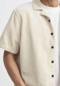 Light beige corduroy shirt with a collared neckline, short sleeves, and six black buttons on the front. Textured fabric with ribbed pattern.