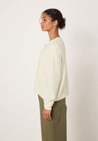 Cream-colored, oversized cardigan in soft fabric with long sleeves, rounded hem, and button closure featuring a simple design.
