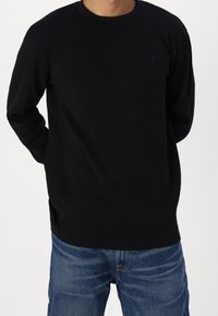 Maglione a maniche lunghe nero a coste con collo tondo e dettagli logo sottili. Abbinato a jeans in denim blu, che mostrano una vestibilità rilassata.