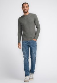 Suéter gris texturizado con cuello redondo, combinado con jeans de color azul claro y zapatillas beige, de pie contra un fondo blanco.