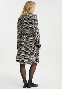 Czarna i beżowa sukienka w wzory z zaznaczoną talią, długimi rękawami i kontrastującymi czarnymi mankietami. Modelka nosi czarne baleriny i przezroczyste rajstopy.