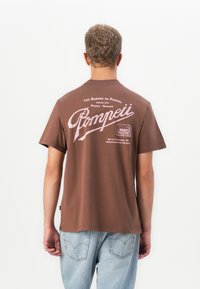 Man wearing brown t-shirt with pink "Pompeii" text and soap graphic on back, paired with light blue jeans, standing against white background.