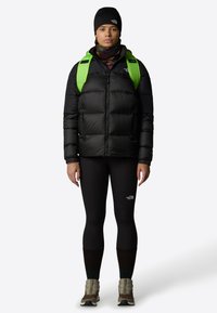 Zwarte pufferjack met een hoge kraag, gecombineerd met zwarte leggings. Heeft een groene rugzak en grijze laarzen. Een muts en nekkraag maken de look compleet.