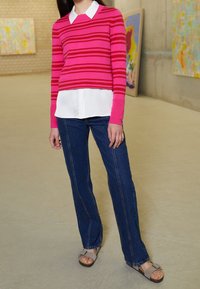 Maglione a righe rosa sopra una camicia bianca, abbinato a jeans in denim blu e sandali grigi. Design semplice, stile aderente, con colori a contrasto.