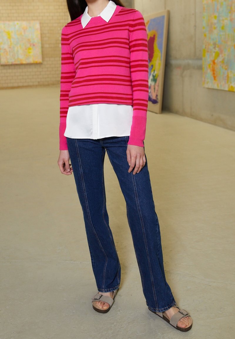 Maglione a righe rosa sopra una camicia bianca, abbinato a jeans in denim blu e sandali grigi. Design semplice, stile aderente, con colori a contrasto.