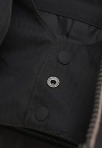 Svart tyginteriör med fiskbensmönster, med en silverfärgad dekorativ tryckknapp och en matt svart knapp. Zipprad kant synlig.