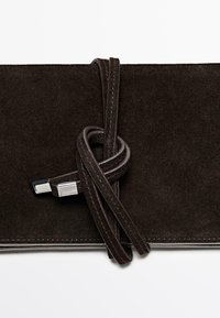 Clutch in camoscio marrone scuro con una tracolla in camoscio avvolgente, con punte in metallo argentato, su sfondo bianco uniforme.
