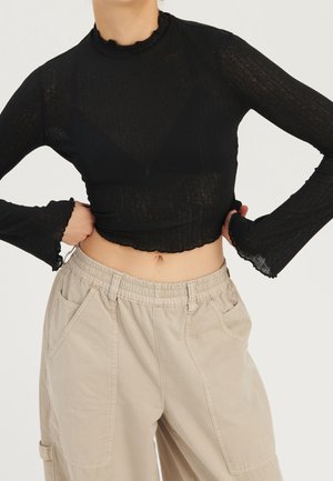 Person trägt ein schwarzes transparentes, langärmliges Cropped-Top mit gerafften Kanten und beige hoch taillierte Cargo-Hosen.