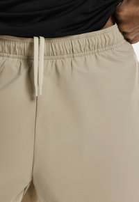 Beigā krāsā šorti ar elastīgu jostu, kurai ir šnorlēdējs, gluda tekstūra un brīvs piegriezums. Melna krekla daļa redzama augšpusē.