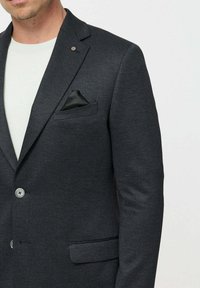 Férfi sötétszürke blézerben, fekete zsebkendővel, fehér kereknyakú póló fölött, nyaktól derékig látható.