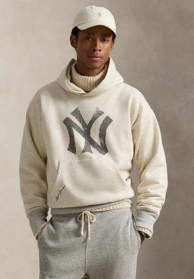 Κρέμα φούτερ με μεγάλο μαύρο λογότυπο NY, συνδυασμένο με γκρι φόρμα που διαθέτει κορδόνι. Το σύνολο έχει χαλαρή εφαρμογή και ρυτιδωτές λεπτομέρειες.