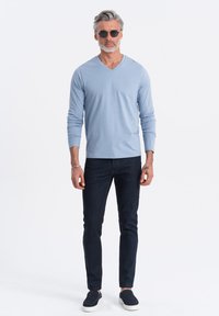 Helles blaues, langärmliges V-Ausschnitt-Shirt kombiniert mit dunklen Jeans und marineblauen Slippern. Das Model trägt runde Sonnenbrillen und hat kurzes graues Haar.