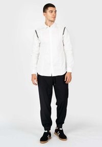 Witte langemouwen shirt met zwarte ritsaccenten op de schouders, gecombineerd met zwarte joggers en zwarte sneakers met witte strepen.