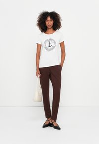 Camiseta branca com um logótipo de âncora redondo, calças ajustadas castanhas, sapatos planos pretos e uma pequena mala de cor clara. Conjunto simples e casual.