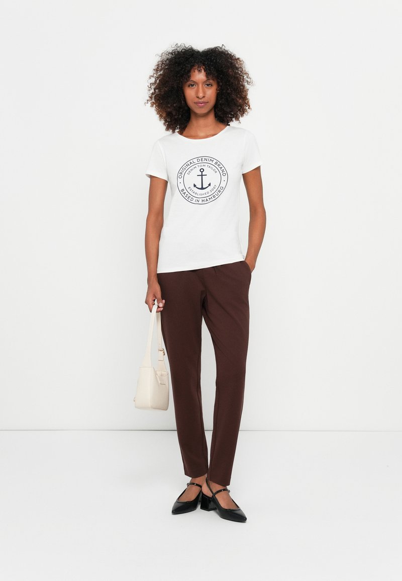 Camiseta branca com um logótipo de âncora redondo, calças ajustadas castanhas, sapatos planos pretos e uma pequena mala de cor clara. Conjunto simples e casual.