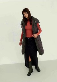 Frau trägt einen langen schwarzen Rock mit Schlitz vorne, einen orangefarbenen gerippten Pullover, eine ärmellose dunkle gepolsterte Weste und schwarze Knöchelstiefel mit Riemen.