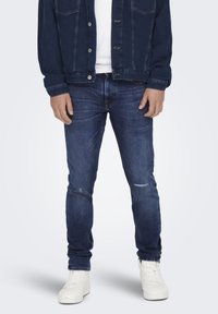 Jachetă din denim închis cu închidere prin nasturi, asociată cu blugi skinny de culoare albastru închis cu accente distruse. Adidașii albi completează outfitul.