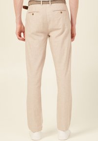 Ljusbeige linnebyxor med rak passform, med bakfickor med knappar och ett texturerat brunt bälte. Vita sneakers synliga.