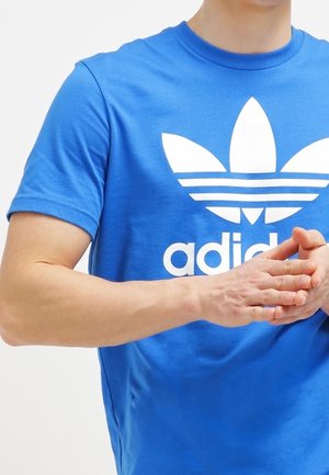 Personne portant un t-shirt Adidas bleu avec logo blanc, mains jointes au niveau de la taille sur un fond blanc uni.