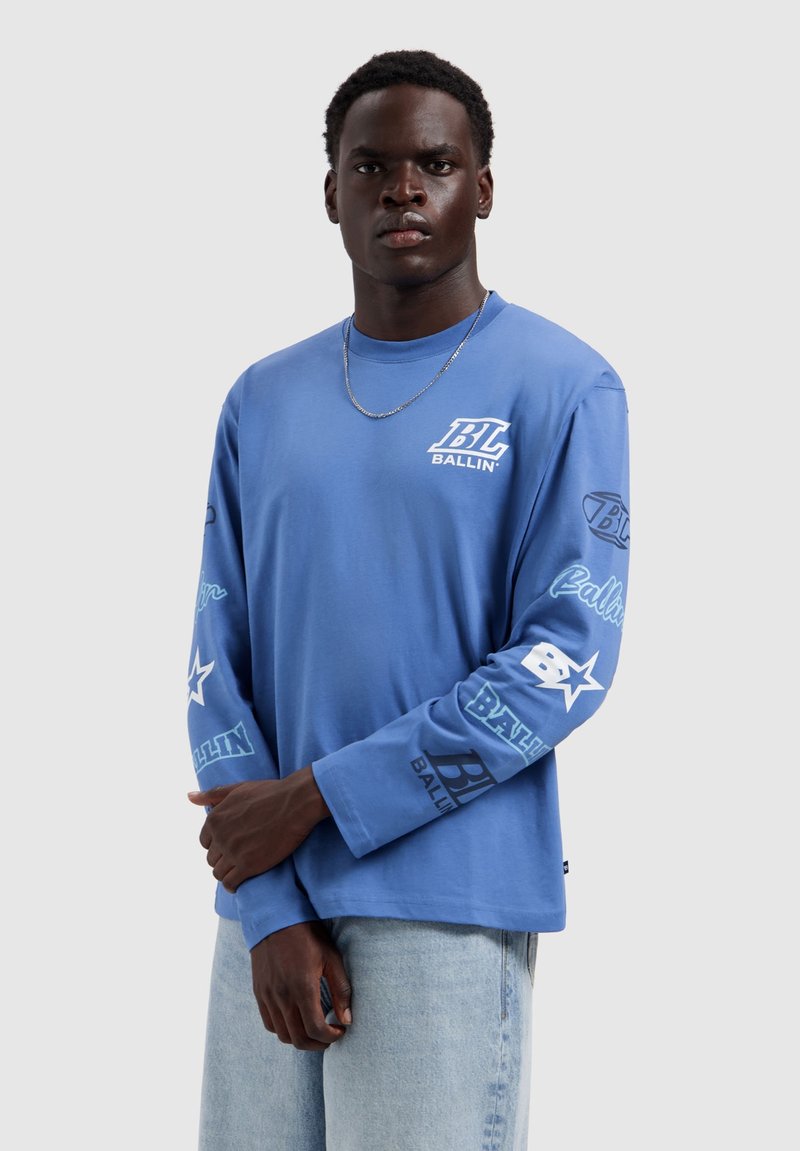 Man draagt een lichtblauw shirt met lange mouwen met "BL Ballin'" logo's op de borst en mouwen, gecombineerd met lichtblauwe spijkerbroeken, tegen een effen achtergrond.