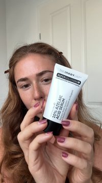 Tubo blanco con tapa negra etiquetado "Serum de Ácido Azelaico al 10%", con texto en negrita y superficie lisa. Sostenido con una mano con uñas de longitud media, esmalte rosa.