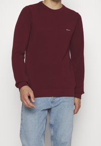 Bordeaux-kleurige katoenen crewneck sweater met ribgebreide manchetten en zoom, voorzien van een klein logo op de borst. Gecombineerd met lichtblauwe denim jeans.