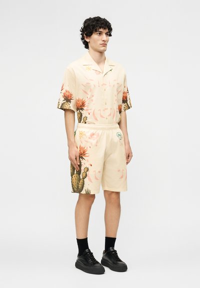 Νεαρός άνδρας που στέκεται, φοράει μια κρεμ κοντομάνικη πουκάμισο και ταιριαστά σορτς με πορτοκαλί σχέδια κάκτων, μαύρες κάλτσες και μαύρα αθλητικά παπούτσια.