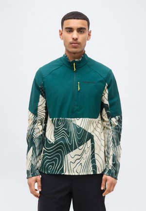 Vyras dėvi žalią ir kreminės spalvos ketvirčio užtrauktuko vilnonį megztinį su topografiniu raštu ir juodas kelnes, stovintis priešais vienspalvį šviesų foną.