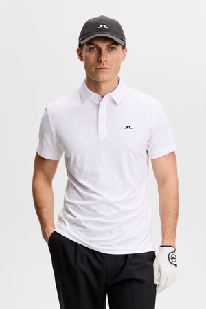 Mand iført en hvid polo, sorte bukser, sort kasket og hvid golfhandske, stående med den ene hånd i lommen mod en hvid baggrund.