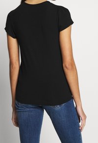 Kvinde iført en ensfarvet sort t-shirt med korte ærmer og blå denimjeans, vist fra ryggen mod en ensartet baggrund.