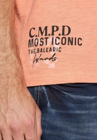 T-shirt w kolorze brzoskwiniowym, wykonany z miękkiego materiału, z czarnym nadrukowanym tekstem i subtelną wizualną fakturą. Noszony z ciemnoniebieskimi dżinsami.