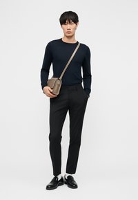 Vīrietis, kurš valkā melnu garroku kreklu, melnas šauras bikses, melnas loaferus ar baltiem zeķēm un bēšu krāsas krustenisku somu, stāv pret vienkāršu balto fonu.