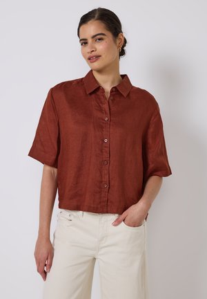 Rust-colored, short-sleeve linen shirt with a pointed collar, a button-down front, and a relaxed fit, paired with white wide-leg pants.