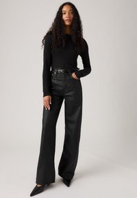 Zwarte lange mouwen top en zwarte hoogzittende wijde jeans met een riem, met een gladde textuur en strakke lijnen. Puntige zwarte hakken.