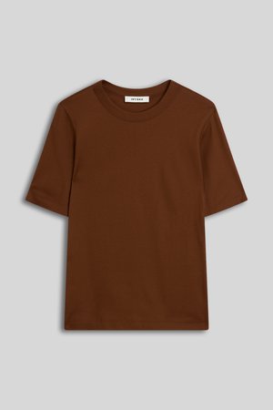 Jeune femme aux cheveux bouclés portant des boucles d'oreilles dorées en forme de créoles, un t-shirt marron ajusté à manches courtes et un pantalon bleu foncé, debout devant un fond uni.