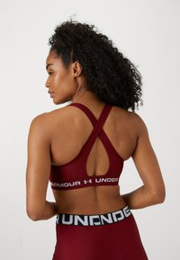 Sostén deportivo en color burdeos con diseño de espalda cruzada, que presenta una banda de logo en blanco. Textura suave y forma ajustada para brindar soporte.