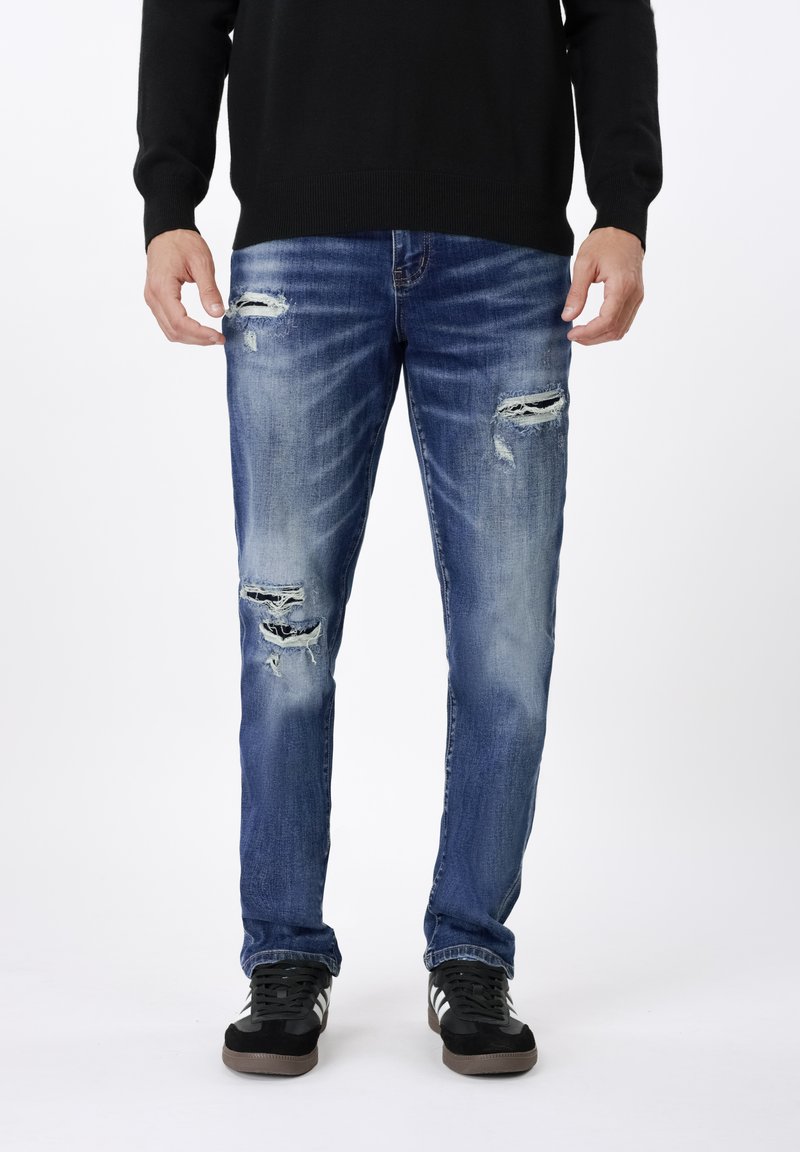 Ξεθωριασμένο μπλε τζιν με σκισίματα στα γόνατα, με ίσια γραμμή στο πόδι, συνδυασμένο με μαύρα αθλητικά παπούτσια για ένα καθημερινό στιλ.