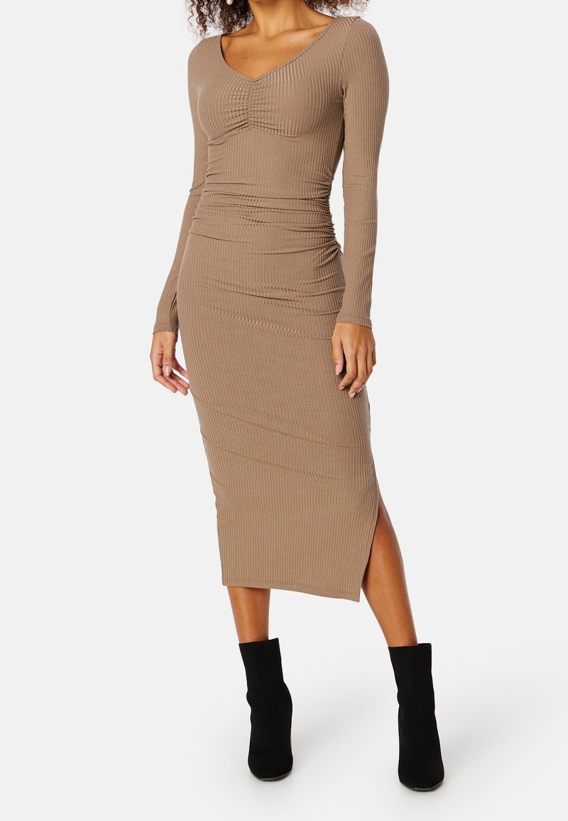 Femme portant une robe midi taupe ajustée, côtelée, à manches longues, avec une fente latérale et des bottines noires, debout sur un fond uni.