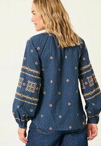 Marineblaue Bluse mit gemustertem Stickereimuster in den Farben Rot, Grün und Gelb. Hat lange Ärmel und einen gerafften Rundhalsausschnitt. Strukturierter Stoff.