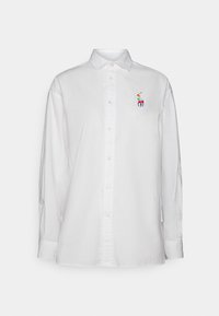 Vit skjorta med knappar, tillverkad av bomull, med långa ärmar, en spetsig krage och en färgglad broderad logotyp på vänster bröst.